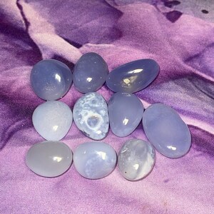 May include: A collection of polished, smooth, blue-gray gemstones of varying shapes and sizes. The stones are arranged on a purple, textured fabric. The gemstones have a translucent quality, with subtle variations in color and pattern.