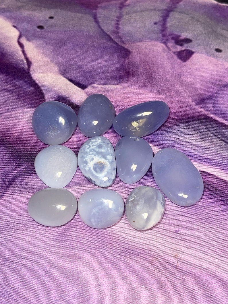 May include: A collection of polished, smooth, blue-gray gemstones of varying shapes and sizes. The stones are arranged on a purple, textured fabric. The gemstones have a translucent quality, with subtle variations in color and pattern.