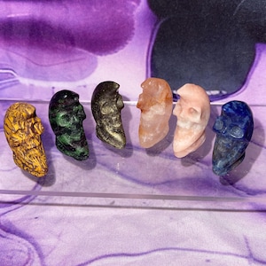 May include: A collection of six carved crystal skulls in various colors, including yellow, green, black, orange, pink, and blue. The skulls are displayed on a clear surface against a purple background.