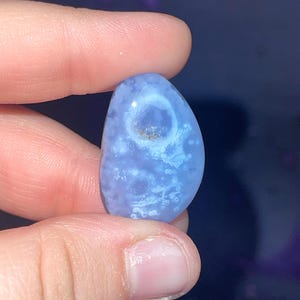 May include: A polished, oval-shaped blue gemstone with white and brown inclusions. The stone is held between fingers, showcasing its smooth texture and natural patterns. The gemstone's color ranges from light to medium blue.
