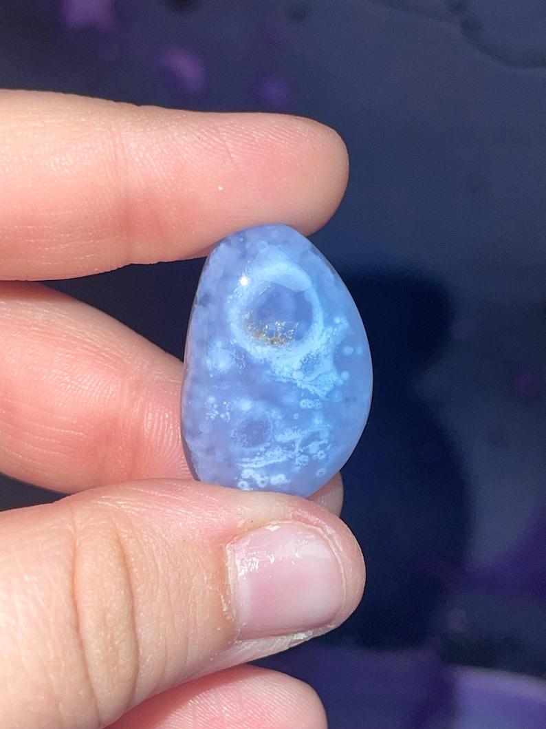 May include: A polished, oval-shaped blue gemstone with white and brown inclusions. The stone is held between fingers, showcasing its smooth texture and natural patterns. The gemstone's color ranges from light to medium blue.