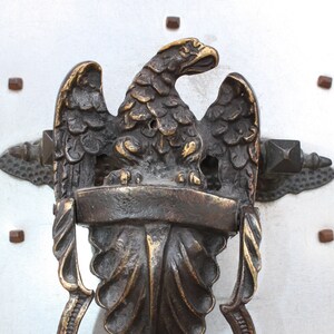 Peut inclure: Un heurtoir de porte en bronze en forme d'aigle avec les ailes déployées. L'aigle tient un bouclier dans ses serres. Le heurtoir est fixé à une porte avec un anneau décoratif.