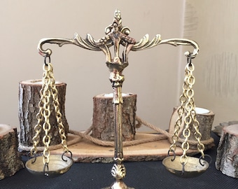 May include: A gold-toned decorative scale with two hanging bowls. The scale is on a wooden surface with a rustic background.