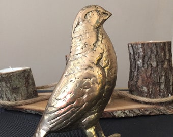 May include: A gold-colored metal bird figurine with detailed wings and feet. The bird is standing on a wooden surface.