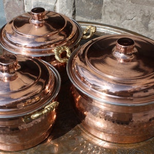 Könnte beinhalten: Drei Kupfertöpfe mit Deckeln und goldenen Griffen. Die Töpfe sind in Pyramidenform auf einem dekorativen Tablett angeordnet.