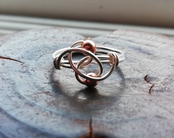 May include: A silver ring with two copper beads, intertwined in a knot design.