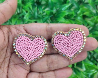 Pode incluir: Dois patches em forma de coração com contas rosa. Cada coração tem uma borda dourada com strass iridescentes. Os corações são preenchidos com pequenas contas rosa. O fundo é verde.