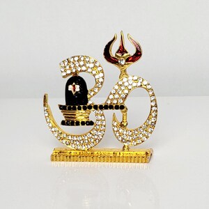 May include: A gold Om symbol with a black and red Shiva Lingam and a trident on top. The Om symbol is decorated with clear rhinestones.