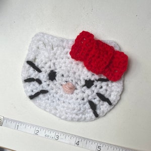 Puede incluir: Una cara de Hello Kitty tejida en blanco con un lazo rojo, bigotes negros y una nariz rosa. El artículo hecho a mano es una pieza decorativa y linda, probablemente un posavasos o una aplicación.