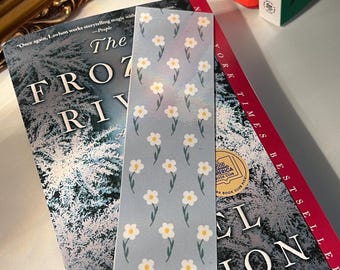 May include: A floral bookmark with a light blue background and a pattern of white daisies with yellow centers. The bookmark is placed on top of a book with the title "The Frozen River".