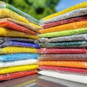 May include: Stacks of small, colorful beads in clear plastic bags. The beads are in various colors, including yellow, red, blue, and silver. The bags are stacked on top of each other, creating a visually appealing display. These beads are ideal for jewelry making and crafts.