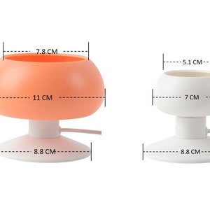 May include: Two suction cup phone holders, one orange and one white. The orange holder is 9 cm tall, 11 cm wide, and 7.8 cm in diameter. The white holder is 8 cm tall, 7 cm wide, and 5.1 cm in diameter. Both holders have a suction cup base that is 8.8 cm in diameter.