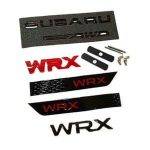 May include: A selection of car emblems and accessories in black and red. Features a textured black emblem with the word "SUBARU" and "AWD", alongside several "WRX" emblems in red and black, plus mounting hardware.