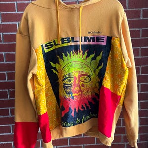 May include: Mustard yellow hoodie with a graphic of a sun with the word "SUBLIME" across the top. The hoodie has red and yellow patterned accents on the sleeves and a "Columbia" logo.