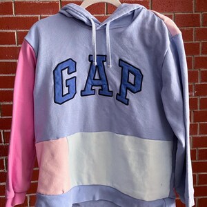 May include: A color-block hoodie in shades of periwinkle, pink, and white. The word "GAP" is embroidered across the chest in a navy blue, outlined in a lighter blue. The hoodie is hanging on a white hanger against a red brick wall.