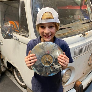 May include: A young person wearing a white baseball cap with a black brim and a blue shirt is holding a silver Volkswagen hubcap clock. The clock has a gold Volkswagen logo and black hands.