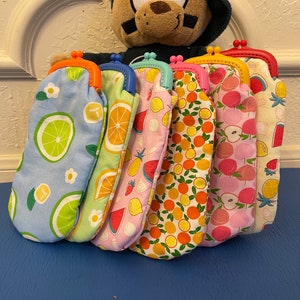 May include: Eight colorful fabric coin purses with different fruit patterns. The purses have a metal clasp and are lined up in a row.