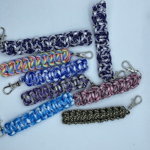 May include: Assortment of colorful paracord keychains with silver key rings. The keychains are made with different patterns and colors, including blue, green, pink, purple, and rainbow.