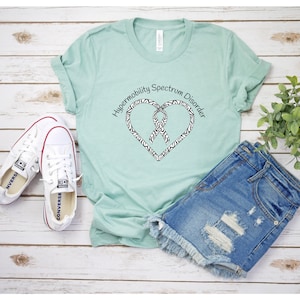 May include: A light green t-shirt with the text "Hypermobility Spectrum Disorder" encircling a heart-shaped design with a zebra print border and a ribbon. The shirt is paired with denim shorts and white sneakers.