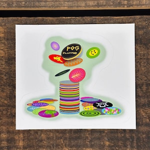 May include: A stack of colorful plastic POGs with a black POG on top that says "POG SLAMMER". There are other POGs scattered around the stack, including a smiley face POG and a POG with a skull and crossbones.