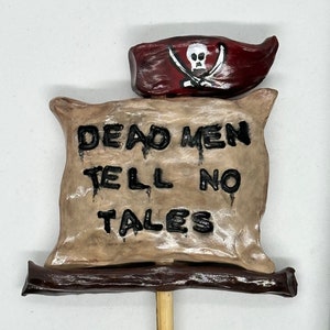May include: A brown paper scroll with the text "Dead Men Tell No Tales" and a red pirate flag with a skull and crossbones on top. The scroll is attached to a wooden stick.