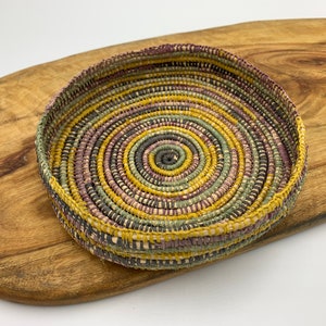 May include: A round woven basket with a spiral pattern in shades of yellow, green, and purple. The basket is made of natural materials and has a rustic, handmade look.