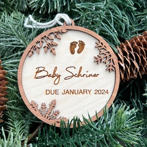 May include: A wooden Christmas ornament with a snowflake design, the text "Baby Schriner DUE JANUARY 2024", and a pair of baby footprints.
