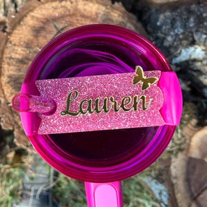 May include: Pink tumbler with a pink glitter name tag that says "Lauren" and a gold butterfly. The tumbler has a clear lid and a straw.