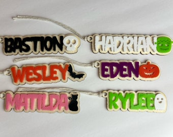 May include: Six wooden Halloween name tags with different names and Halloween-themed icons. The names are Bastion, Hadrian, Wesley, Eden, Matilda, and Rylee. The icons are a skull, a monster, a bat, a pumpkin, a cat, and a ghost.