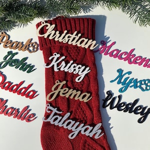 May include: A red knitted stocking with various wooden ornaments with names written on them. The names include: Pearl, John, Dadda, Charlie, Christian, Mackenzie, Krissy, Jema, Talayah, Nyx, and Wesley.