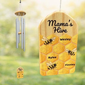 May include: A wooden wind chime with a decorative sign that reads "Mama's Hive". The sign features a honeycomb design with bee illustrations and the names Wesley, Rylee, and Finnley. The wind chime has silver tubes.