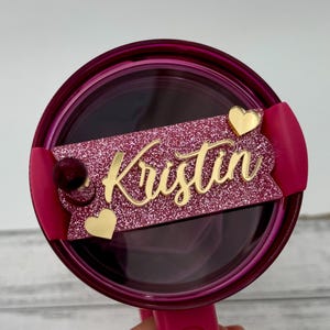 May include: A pink glitter tumbler with a gold nameplate that reads "Kristin." The nameplate is decorated with gold hearts and a small, dark red pom-pom. The tumbler is a vibrant pink color.