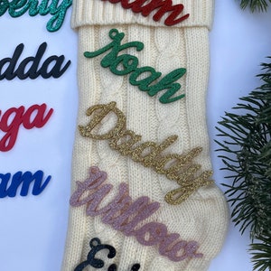 May include: A white knitted stocking with six glittery names on it: Liam, Noah, Daddy, Willow, Erica. The names are in different colors, including red, green, gold, pink, and black.