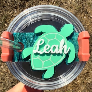 May include: A teal green turtle-shaped tumbler lid with the name "Leah" written in white. The lid is attached to a clear tumbler with a pink straw.