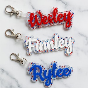 May include: Three personalized name keychains. Each keychain features a different name: "Wesley" in red, "Finley" in white, and "Rylee" in blue. The names are set against a clear background with red and blue star accents, and each has a silver clasp.