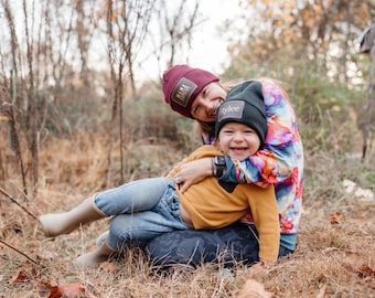 May include: A woman and child wearing beanies with patches. The woman's beanie says "Mama" and the child's says "Rylee". The child is wearing a mustard yellow sweater and blue jeans. The woman is wearing a colorful tie-dye shirt.