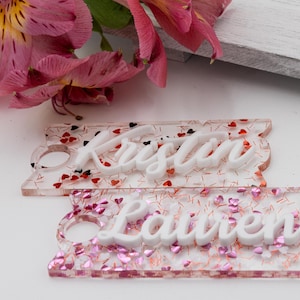 May include: Two clear rectangular keychains with glitter and pink and red hearts. The keychains have the names "Kristen" and "Lauren" written in white raised letters.