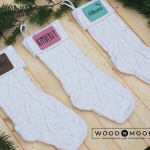 May include: Three white knitted Christmas stockings with personalized name tags. The names on the tags are Rylee, Kimberly, and Athena. The stockings are on a wooden surface.