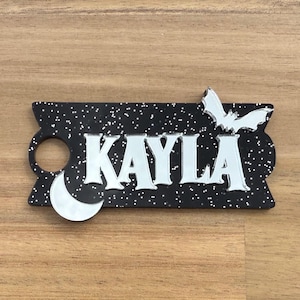 May include: A black name tag with the name "KAYLA" in silver letters. The tag features a crescent moon and a bat, with a starry background. The tag is designed with a Halloween or gothic theme.