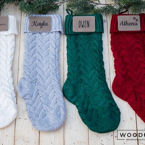 May include: Four knitted Christmas stockings with personalized leather name tags. The stockings are white, light gray, dark green, and red. The names on the tags are Megan, Kayla, Owen, and Athena.