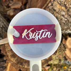 May include: A close-up of a white tumbler lid with a burgundy name tag that says "Kristin" in silver lettering. The name tag is attached to the lid with a clear adhesive.