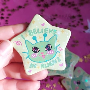 May include: A star-shaped badge featuring a light green alien cat design with the text "I BELIEVE IN ALIENS". The alien cat has large eyes and antennae. The background is a pink surface with gold star-shaped confetti.