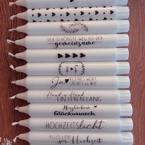 May include: A stack of white candles with black text and heart designs. The candles have messages such as "Verliebt, Verlobt, Verheiratet", "Der sch&ouml;nste Weg ist der gemeinsame", "Ja", "Kleines Wort, Gro&szlig;es Gl&uuml;ck", "Hand in Hand, Ein Leben Lang", "Herzlichen Gl&uuml;ckwunsch", "Hochzeitslicht", and "Alles Liebe zur Hochzeit".