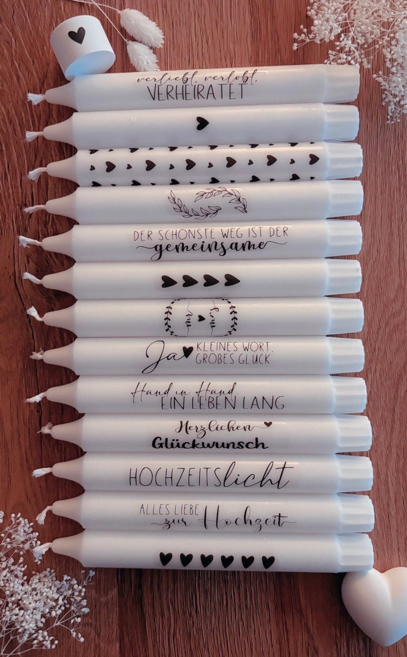 May include: A stack of white candles with black text and heart designs. The candles have messages such as "Verliebt, Verlobt, Verheiratet", "Der sch&ouml;nste Weg ist der gemeinsame", "Ja", "Kleines Wort, Gro&szlig;es Gl&uuml;ck", "Hand in Hand, Ein Leben Lang", "Herzlichen Gl&uuml;ckwunsch", "Hochzeitslicht", and "Alles Liebe zur Hochzeit".