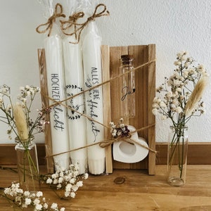 May include: Three white candles wrapped in twine and labeled "Hochzeitslicht" and "Alles Liebe zur Hochzeit". A small wooden box with a heart-shaped candle holder and a glass vial with a cork top. The box is tied with twine and decorated with small white flowers.
