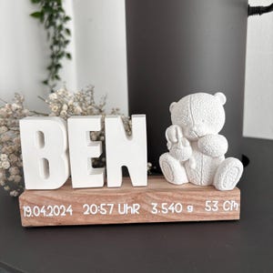 Könnte beinhalten: Ein weißes Dekorationsobjekt mit dem Namen "BEN" in großen Blockbuchstaben. Eine kleine weiße Teddybärfigur sitzt neben den Buchstaben. Die Holzbasis zeigt Datum, Uhrzeit, Gewicht (3.540 g) und Größe (53 cm).