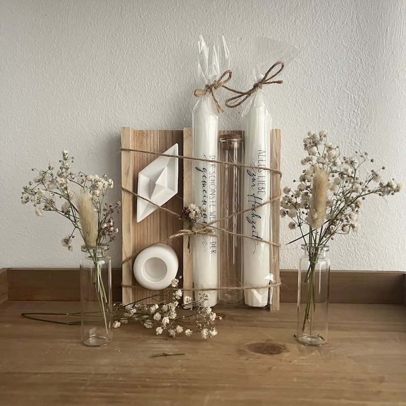 May include: A wooden box with two white candles, a small white boat, and two glass vases with dried flowers. The candles are wrapped in twine and have text that reads "Der sch&ouml;nste Weg" and "Alles Liebe - Gl&uuml;ckzeit".