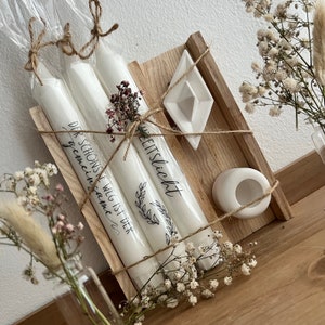May include: Three white candles wrapped in twine and paper with the text "Der Schons Weil ist der gemeinsame" and "Zeitslicht" on them. The candles are in a wooden box with a white ceramic dish and a white ceramic candle holder.