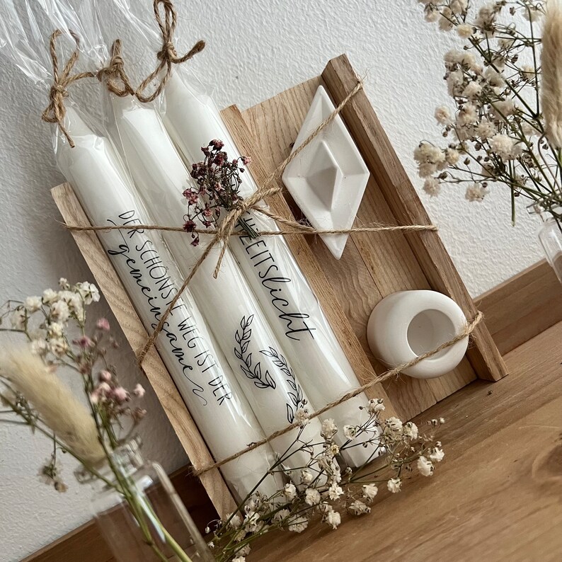 May include: Three white candles wrapped in twine and paper with the text "Der Schons Weil ist der gemeinsame" and "Zeitslicht" on them. The candles are in a wooden box with a white ceramic dish and a white ceramic candle holder.