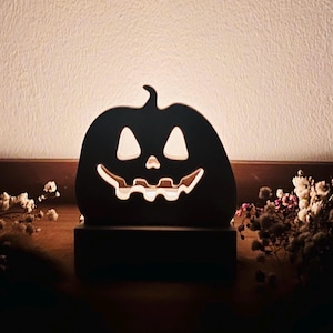 Könnte beinhalten: Eine dunkle Kürbislaterne im Silhouetten-Stil mit einem ausgeschnittenen Jack-o'-Lantern-Gesicht mit dreieckigen Augen und einem gezackten Mund. Der Kürbis ist auf einem rechteckigen Sockel montiert und strahlt ein warmes, sanftes Licht aus.